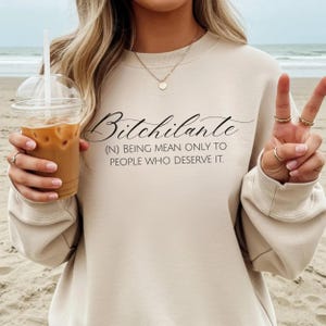May include: A beige sweatshirt with the word "Bitchilante" in a cursive font, followed by the definition: "(N) BEING MEAN ONLY TO PEOPLE WHO DESERVE IT." The person is holding a clear plastic cup with iced coffee.