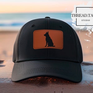 May include: Black baseball cap with a brown leather patch featuring a silhouette of a German Shepherd dog. The patch is stitched onto the front of the cap. The cap has a curved brim and an adjustable strap.