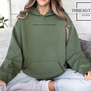 May include: An olive green hoodie with the phrase "coffee is my love language." The hoodie features a front pocket and a drawstring hood. The model is wearing light wash jeans and grey socks.