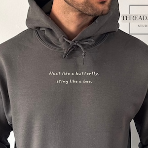 以下が含まれることがあります： 前面に「float like a butterfly, sting like a bee.」という白い文字がプリントされたダークグレーのフード付きスウェットシャツです。