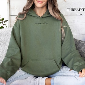 May include: An olive green hoodie with a front pocket and drawstring hood. The text "anxiety-ridden." is printed in black on the upper left chest. The brand name "THREAD.TALKS STUDIO" is printed in black on the upper right.
