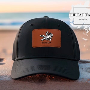 May include: Black baseball cap with a brown leather patch featuring a cartoon astronaut and the words "Spaced Out." The cap has a curved brim and is set against a beach background. The brand name "THREAD.TALKS STUDIO" is also visible.
