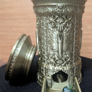 Op de afbeelding: Een gouden metalen dobbelsteentoren met een sierlijk ontwerp. De toren heeft een lade die opengaat om een ​​groene en blauwe dobbelsteen te onthullen. De toren is ontworpen om een ​​blikje Summer Shandy te bevatten.