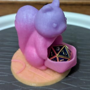 May include: A pink and purple squirrel figurine holding a multi-sided die. The squirrel is on a wooden-looking base. The die is black with gold trim and white numbers. The figurine is likely for a tabletop game.