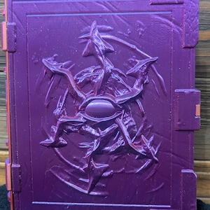 Puede incluir: Un libro morado con un diseño en relieve de una estrella de cinco puntas con un ojo en el centro.