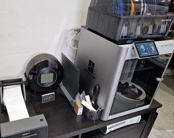 May include: A black and silver 3D printer with a clear filament holder on top. The printer is on a black table with a black and white printer, a black speaker, and a black scale.