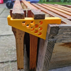 Op de afbeelding: Een gele plastic bijenkorf-raamspatie met een honingraatpatroon. De spatie is bevestigd aan een houten bijenkorf-raam.