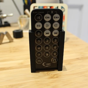 Può includere: Un telecomando nero con una varietà di pulsanti, tra cui play/pausa, canale su/giù e tasti numerici. Il telecomando è in un supporto di plastica nero. Il testo "car-mel" è stampato sul telecomando.