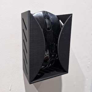 May include: A black 3D printed wall mount for a computer mouse. The mount has a curved design to hold the mouse securely.