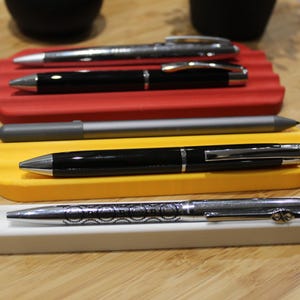 May include: Four pens are displayed in a row, each resting in a separate holder. The holders are made of different colours: red, yellow, and white. The pens are silver, black, and grey.