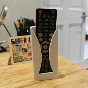 Puede incluir: Soporte para mando a distancia blanco impreso en 3D con un mando a distancia negro en su interior. El soporte tiene un diseño curvo y está sobre una superficie de madera.