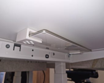 Puede incluir: Un soporte de montaje de plástico blanco fijado en la parte inferior de una superficie blanca, que sostiene un dispositivo electrónico fino y rectangular de color plateado y blanco. El soporte tiene una pequeña rejilla de muescas circulares.