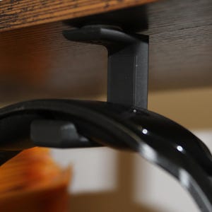 Puede incluir: Gancho negro para auriculares montado debajo de un estante de madera. El gancho está hecho de plástico y tiene un diseño simple. Los auriculares son negros y tienen una forma curva.