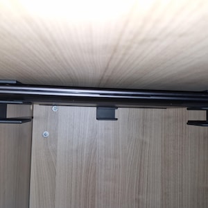 Op de afbeelding: Een laptopcomputer is met een zwarte metalen beugel aan een muur gemonteerd. De laptop is gesloten en de beugel is met schroeven aan de muur bevestigd.