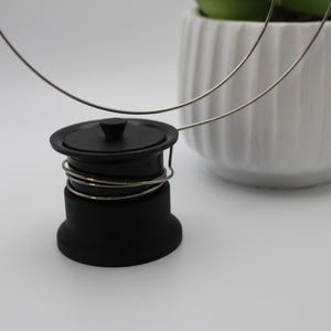 Könnte beinhalten: Ein schwarzer Spülbeckenstöpsel mit silbernen Drahtdetails. Der Stöpsel befindet sich neben einem weißen Keramiktopf mit einer grünen Pflanze. Der Hintergrund ist eine schlichte weiße Oberfläche.