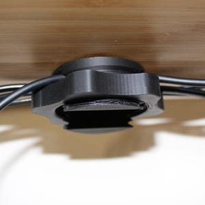 May include: A black plastic cable management clip with multiple wires running through it. The clip is attached to a wooden surface.