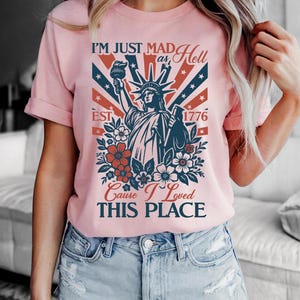May include: Pale pink t-shirt with a graphic of the Statue of Liberty, text reading "I'M JUST MAD as Hell Cause I Loved THIS PLACE" and "EST 1776". The design incorporates red, white, and blue elements and floral details.