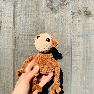 K&ouml;nnte beinhalten: Eine handgefertigte H&auml;kelpuppe in Form eines Affen mit hellbraunem Kopf und braunem K&ouml;rper, Armen und Beinen. Der Affe hat schwarze Augen und ist aus weichem Garn gefertigt. Die Puppe wird in einer Hand vor einem Holzgrund gehalten.