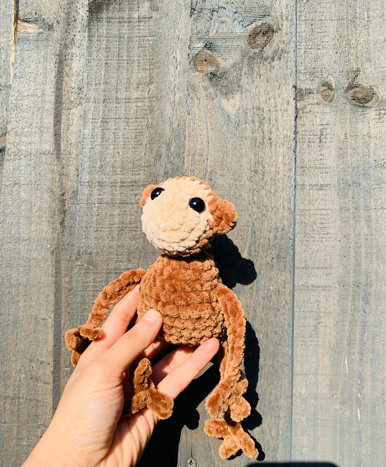 K&ouml;nnte beinhalten: Eine handgefertigte H&auml;kelpuppe in Form eines Affen mit hellbraunem Kopf und braunem K&ouml;rper, Armen und Beinen. Der Affe hat schwarze Augen und ist aus weichem Garn gefertigt. Die Puppe wird in einer Hand vor einem Holzgrund gehalten.