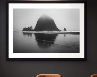 May include: A black and white framed landscape photograph depicting a large sea stack rising from the ocean, with its reflection visible on the wet sandy beach. The scene is shrouded in thick fog, creating a moody atmosphere with soft light.