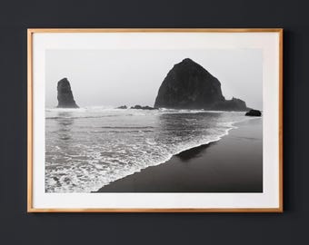 May include: A black and white framed photograph depicting a coastal scene with two large rock formations rising from the ocean. The foreground shows wet sand and gentle waves washing onto the shore under a hazy, overcast sky in a wooden frame.