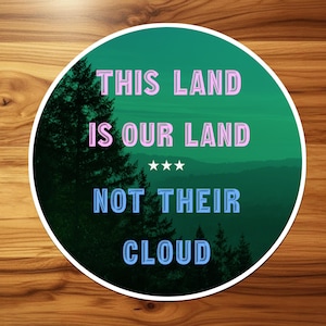 Op de afbeelding: Een ronde sticker met een donkergroen bos- en berglandschap. De tekst luidt THIS LAND IS OUR LAND in roze letters, gevolgd door drie witte sterren en NOT THEIR CLOUD in lichtblauwe letters met een witte rand.