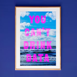 Op de afbeelding: Een ingelijste kunstdruk met een foto van een blauwe oceaan en lucht met witte wolken. De tekst YOU CAN'T DRINK DATA is in heldere neonroze hoofdletters in het midden van de afbeelding gedrukt, over de horizonlijn heen.