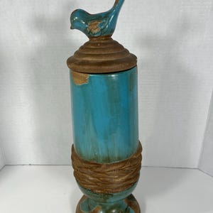 May include: A tall, cylindrical ceramic canister featuring a vibrant turquoise glazed finish with distressed brown accents. The lid is topped with a sculpted turquoise bird figurine, and the base is wrapped in a textured, brown, rope-like design.