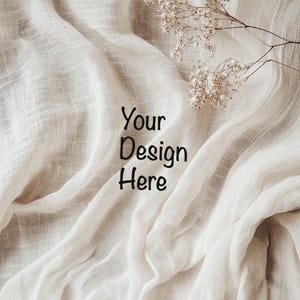 Puede incluir: Un telón de fondo de tela blanca texturizada sobre un fondo marrón claro. La tela está drapeada y doblada, creando una sensación suave y aireada. El texto "Your Design Here" está escrito en negro sobre la tela.