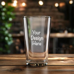 May include: A clear glass pint glass with the text "Your Design Here" printed on the side. The glass is sitting on a wooden table.