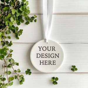 May include: White ceramic ornament with a white ribbon hanging from it. The ornament has the text "YOUR DESIGN HERE" printed on it. The ornament is on a white wooden background with green leaves.