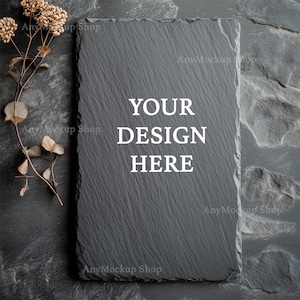 Peut inclure: Une planche à découper en ardoise noire avec le texte "YOUR DESIGN HERE" en blanc. La planche est posée sur une surface texturée gris foncé avec une brindille florale séchée.