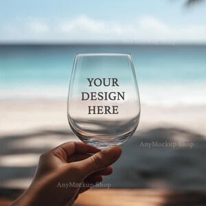May include: A clear glass wine glass with the text "YOUR DESIGN HERE" printed on the side. The glass is being held by a hand against a blurred background of a beach and ocean.