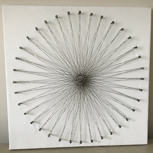 Puede incluir: Un lienzo blanco con un diseño circular creado por una cuerda estirada entre clavos. La cuerda está tejida en un patrón radial, creando un efecto de estrella.