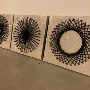 Könnte beinhalten: Drei quadratische Leinwände mit schwarzen String-Art-Designs. Jede Leinwand zeigt ein anderes geometrisches Muster, wobei ein Design einen Kreis in einem Kreis zeigt.