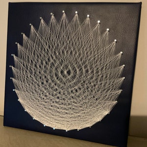 May include: White string art on a dark blue background. The string is stretched between nails in a circular pattern, creating a geometric design.