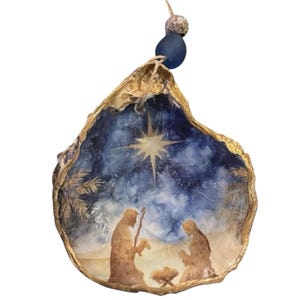 May include: A decorative oyster shell ornament featuring a painted nativity scene with a bright star against a dark blue night sky. The shell edges are gilded in gold, and it hangs from a string adorned with a blue bead and a smaller speckled bead.