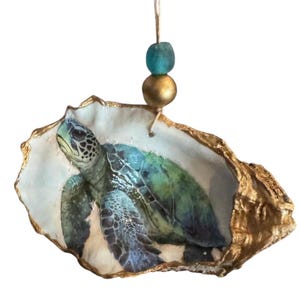 May include: A decorative oyster shell ornament featuring a hand-painted sea turtle in shades of green and blue. The shell edges are gilded in gold, and it hangs from a twine string adorned with one teal bead and one metallic gold bead.