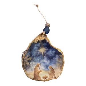 May include: Hand-painted oyster shell Christmas ornament. The shell has a gold-painted edge and features a nativity scene with a star in a night sky. A blue bead and string are attached for hanging.