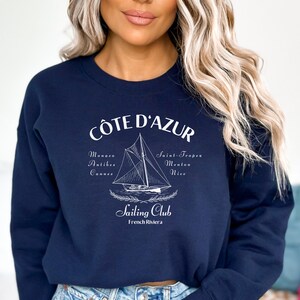 Peut inclure: Sweat-shirt bleu marine avec un motif blanc. Le motif indique "Cote D'Azur Sailing Club French Riviera" avec un voilier et les noms des villes de la Côte d'Azur. Le sweat-shirt est de style col rond.
