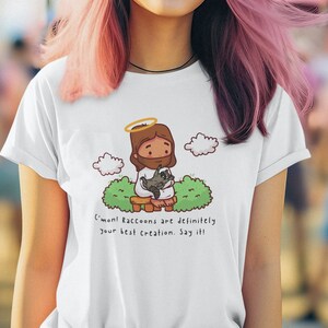 May include: A white t-shirt with a cartoon illustration of Jesus holding a raccoon. The text on the shirt reads "C'mon! Raccoons are definitely your best creation. Say it!"