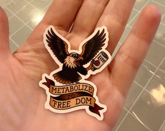 May include: A sticker featuring a bald eagle with outstretched wings, holding a can of beer. The eagle is brown, black, and white, with a red beak. A banner below reads "Metabolize to Free Dom."