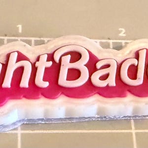 May include: A white and pink Flight Baddie sticker. The text "Flight Baddie" is in white, with a pink background and a white border. The sticker is on a gray surface with measurement markings.