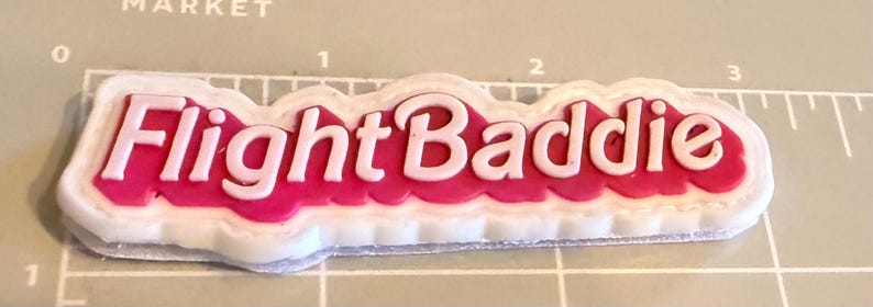 May include: A white and pink Flight Baddie sticker. The text "Flight Baddie" is in white, with a pink background and a white border. The sticker is on a gray surface with measurement markings.