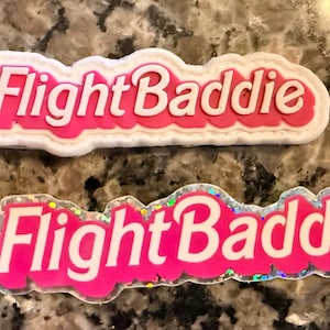 May include: Two pink shoe charms with the text "Flight Baddie". The charms have white and iridescent details. One charm has a white border, and the other has a glittery border.