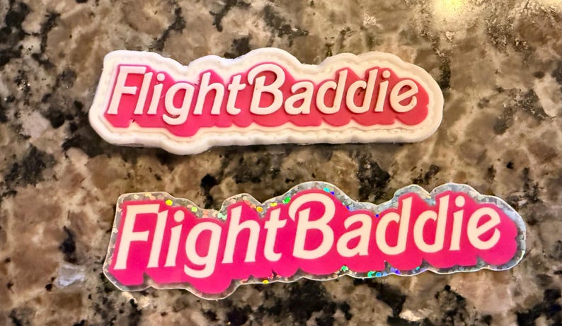 May include: Two pink shoe charms with the text "Flight Baddie". The charms have white and iridescent details. One charm has a white border, and the other has a glittery border.