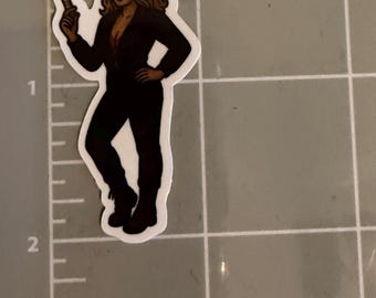 May include: A sticker featuring a woman in a black jumpsuit, white helmet with a red cross, and holding a syringe. The woman has brown hair and is wearing a visor. The sticker is on a gray surface with measurement markings.