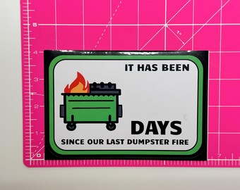 May include: A rectangular sticker with a white background and a green border. It features a green dumpster with orange and red flames, and the black text: IT HAS BEEN DAYS SINCE OUR LAST DUMPSTER FIRE. The sticker is placed on a pink cutting mat.