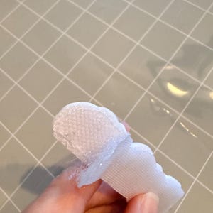 May include: A white, scalloped-edge fabric piece with a textured surface, possibly a decorative element or trim. The fabric is held against a light grey grid surface.