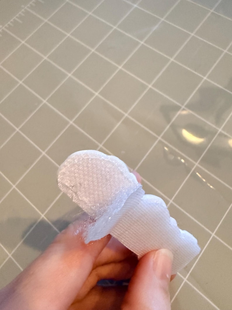 May include: A white, scalloped-edge fabric piece with a textured surface, possibly a decorative element or trim. The fabric is held against a light grey grid surface.